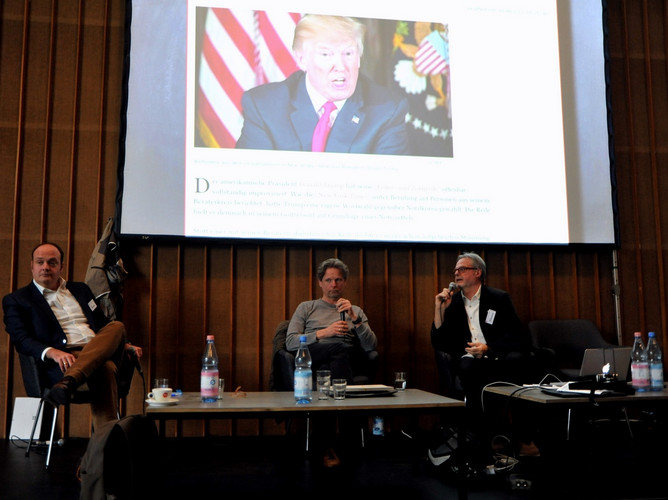 Vortrag mit drei Personen auf Bühne mit Bild von Donald Trump.