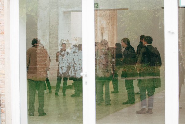 People in an exhibition room photographed through a reflecting glass door.