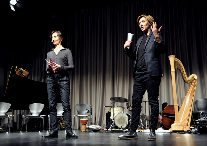 Two People on stage with microfone in front of Musical Instruments.