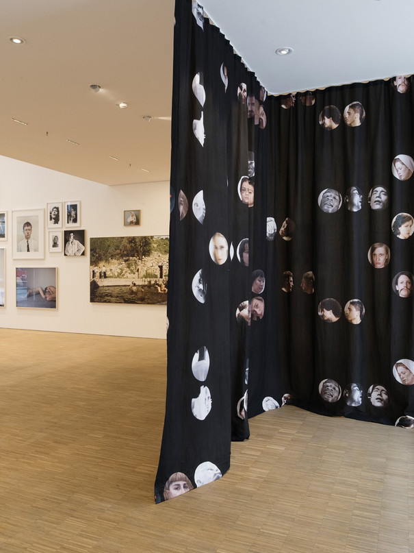 Blick in die Ausstellung, ein schwarzer Vorhang mit Gesichtern teilt den Raum