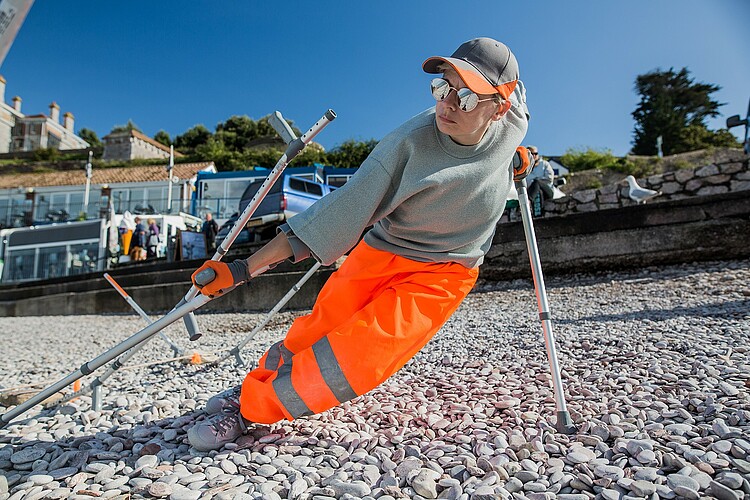 Facing the camera Claire is in a deep diagonal leaning position towards the right of the image, her weight held by her left crutch slightly behind her.  Her legs and other crutch  are stretched out to the right hand side. And she is looking towards them.  Parts of other crutches stick up out of the pebbles on the beach behind her.