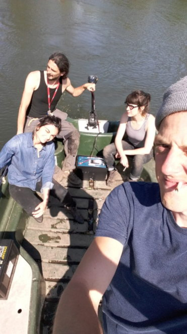 Ein junger Mann mit Mütze und Kippe macht ein Selfie mit 3 Freunden im Hintergund auf einem Floß.