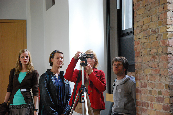 Eine Gruppe Studenten steht hinter einem Stativ, auf dem eine Videokamera montiert ist.