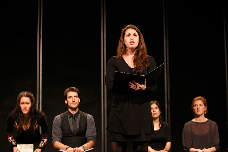 Eine Sängerin steht auf der Bühne, hinter ihr vier sitzende Studierende